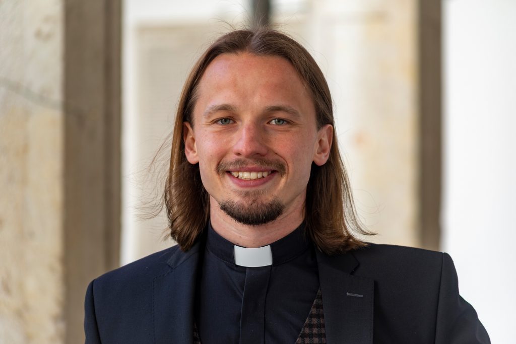 Ein lächelnder Priester in Anzug und mit Kragen steht im Licht einer Kirche, die zur Pfarrei Seligenporten gehört.